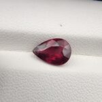 High-resolution macro photograph of a 2.71-carat natural Madagascar Rhodolite Garnet gemstone resting on a white textured leather display bed, showcasing a masterfully faceted pear cut with rich, saturated raspberry-red hues and brilliant purplish-pink fire.