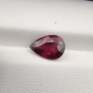 High-resolution macro photograph of a 2.71-carat natural Madagascar Rhodolite Garnet gemstone resting on a white textured leather display bed, showcasing a masterfully faceted pear cut with rich, saturated raspberry-red hues and brilliant purplish-pink fire.