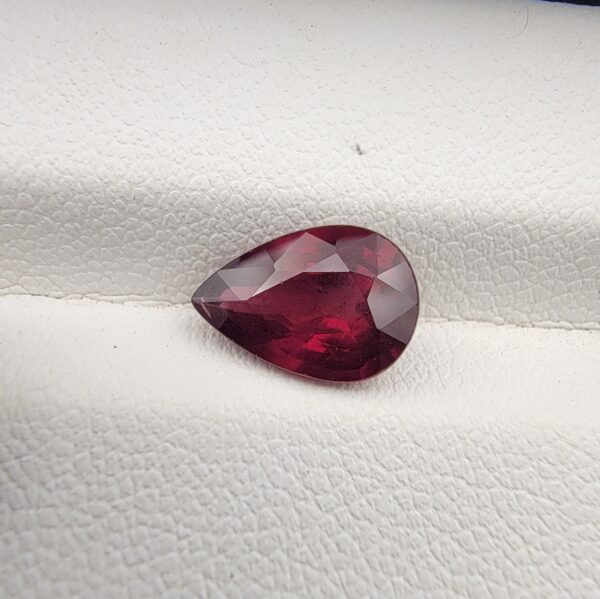 High-resolution macro photograph of a 2.71-carat natural Madagascar Rhodolite Garnet gemstone resting on a white textured leather display bed, showcasing a masterfully faceted pear cut with rich, saturated raspberry-red hues and brilliant purplish-pink fire.