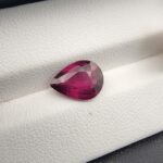 High-resolution macro photograph of a 3.02-carat natural Madagascar Rhodolite Garnet gemstone resting on a white textured leather display bed, showcasing a masterfully faceted pear cut with rich, saturated raspberry-red hues and brilliant purplish-pink fire.