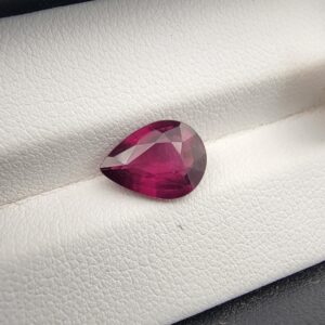 High-resolution macro photograph of a 3.02-carat natural Madagascar Rhodolite Garnet gemstone resting on a white textured leather display bed, showcasing a masterfully faceted pear cut with rich, saturated raspberry-red hues and brilliant purplish-pink fire.