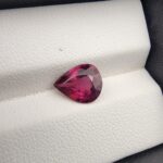 High-resolution macro photograph of a 2.29-carat natural Madagascar Rhodolite Garnet gemstone set against a white textured leather display cushion, displaying a brilliantly faceted pear cut with rich, saturated raspberry-red coloration and deep purplish-pink flashes.