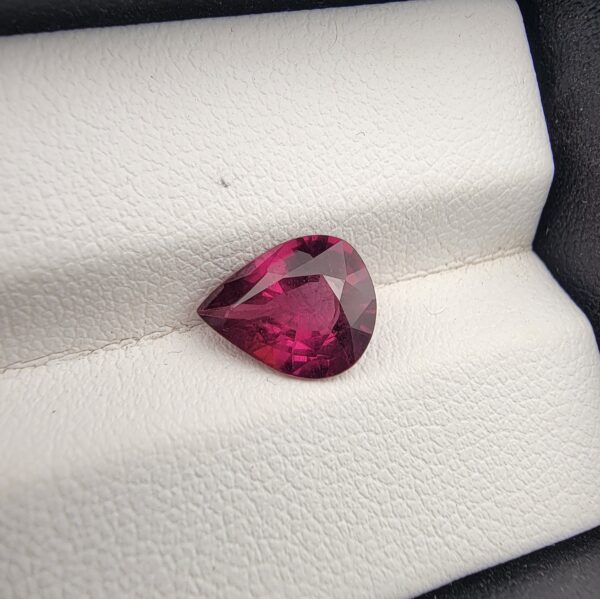 High-resolution macro photograph of a 2.29-carat natural Madagascar Rhodolite Garnet gemstone set against a white textured leather display cushion, displaying a brilliantly faceted pear cut with rich, saturated raspberry-red coloration and deep purplish-pink flashes.