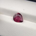 High-resolution macro photograph of a 1.98-carat natural Madagascar Rhodolite Garnet gemstone resting on a white textured leather display bed, showcasing a masterfully faceted pear cut with rich, saturated raspberry-red hues and brilliant purplish-pink fire.