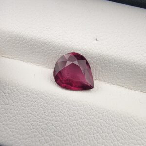High-resolution macro photograph of a 1.98-carat natural Madagascar Rhodolite Garnet gemstone resting on a white textured leather display bed, showcasing a masterfully faceted pear cut with rich, saturated raspberry-red hues and brilliant purplish-pink fire.