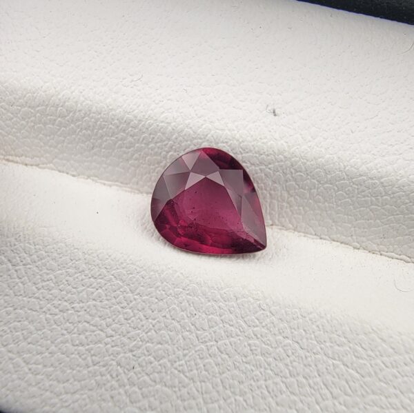 High-resolution macro photograph of a 1.98-carat natural Madagascar Rhodolite Garnet gemstone resting on a white textured leather display bed, showcasing a masterfully faceted pear cut with rich, saturated raspberry-red hues and brilliant purplish-pink fire.