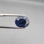 Macro photograph of an 8.09-carat natural unheated Afghan blue indigo sapphire (Neelam) held in stainless steel gem tweezers against a neutral grey backdrop, showcasing a faceted oval cut, deep indigo blue body color, and natural silk inclusions.