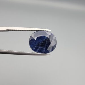 Macro photograph of an 8.09-carat natural unheated Afghan blue indigo sapphire (Neelam) held in stainless steel gem tweezers against a neutral grey backdrop, showcasing a faceted oval cut, deep indigo blue body color, and natural silk inclusions.