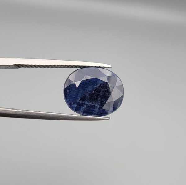 Macro photograph of an 8.09-carat natural unheated Afghan blue indigo sapphire (Neelam) held in stainless steel gem tweezers against a neutral grey backdrop, showcasing a faceted oval cut, deep indigo blue body color, and natural silk inclusions.