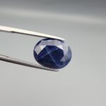 ​Multi-angle macro photographs of a massive 13.10-carat natural unheated Afghan blue indigo sapphire (Neelam) held in stainless steel gem tweezers against a neutral grey background, displaying a faceted oval cut, deep indigo blue saturation, and delicate silk inclusion patterning.