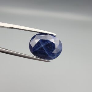 ​Multi-angle macro photographs of a massive 13.10-carat natural unheated Afghan blue indigo sapphire (Neelam) held in stainless steel gem tweezers against a neutral grey background, displaying a faceted oval cut, deep indigo blue saturation, and delicate silk inclusion patterning.
