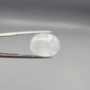 Macro photograph of a 15.30-carat natural Durr-e-Najaf (Clear Rock Crystal Quartz) cabochon held in metal gem tweezers against a neutral backdrop, displaying an oval polished cut, pure water-like transparency, and delicate natural internal veil inclusions.