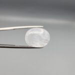 Macro photograph of a 15.30-carat natural Durr-e-Najaf (Clear Rock Crystal Quartz) cabochon held in metal gem tweezers against a neutral backdrop, displaying an oval polished cut, pure water-like transparency, and delicate natural internal veil inclusions.