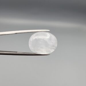 Macro photograph of a 15.30-carat natural Durr-e-Najaf (Clear Rock Crystal Quartz) cabochon held in metal gem tweezers against a neutral backdrop, displaying an oval polished cut, pure water-like transparency, and delicate natural internal veil inclusions.