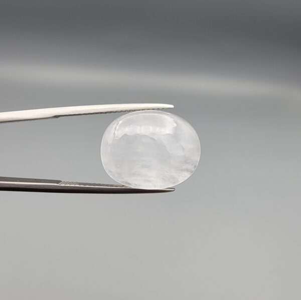 Macro photograph of a 15.30-carat natural Durr-e-Najaf (Clear Rock Crystal Quartz) cabochon held in metal gem tweezers against a neutral backdrop, displaying an oval polished cut, pure water-like transparency, and delicate natural internal veil inclusions.