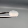 Macro photograph of a 15.30-carat natural Durr-e-Najaf (Clear Rock Crystal Quartz) cabochon held in metal gem tweezers against a neutral backdrop, displaying an oval polished cut, pure water-like transparency, and delicate natural internal veil inclusions.