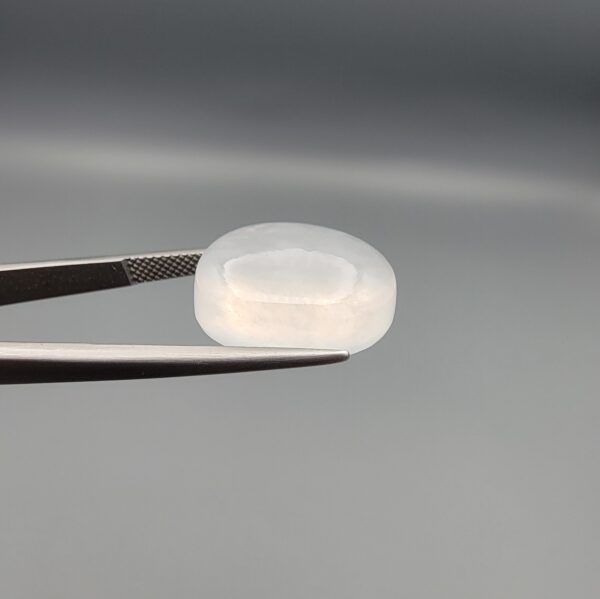 Macro photograph of a 15.30-carat natural Durr-e-Najaf (Clear Rock Crystal Quartz) cabochon held in metal gem tweezers against a neutral backdrop, displaying an oval polished cut, pure water-like transparency, and delicate natural internal veil inclusions.