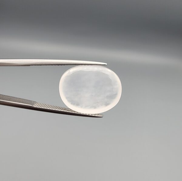 Macro photograph of a 15.30-carat natural Durr-e-Najaf (Clear Rock Crystal Quartz) cabochon held in metal gem tweezers against a neutral backdrop, displaying an oval polished cut, pure water-like transparency, and delicate natural internal veil inclusions.
