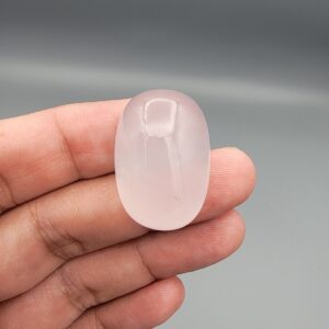 Macro photograph showing a monumental 75.44-carat natural Durr-e-Najaf (Clear Rock Crystal Quartz) cabochon resting on fingertips, featuring a large smooth oval dome, water-clear transparency, and subtle organic internal veil inclusions.