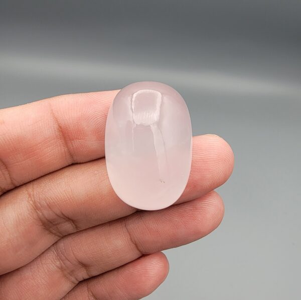 Macro photograph showing a monumental 75.44-carat natural Durr-e-Najaf (Clear Rock Crystal Quartz) cabochon resting on fingertips, featuring a large smooth oval dome, water-clear transparency, and subtle organic internal veil inclusions.