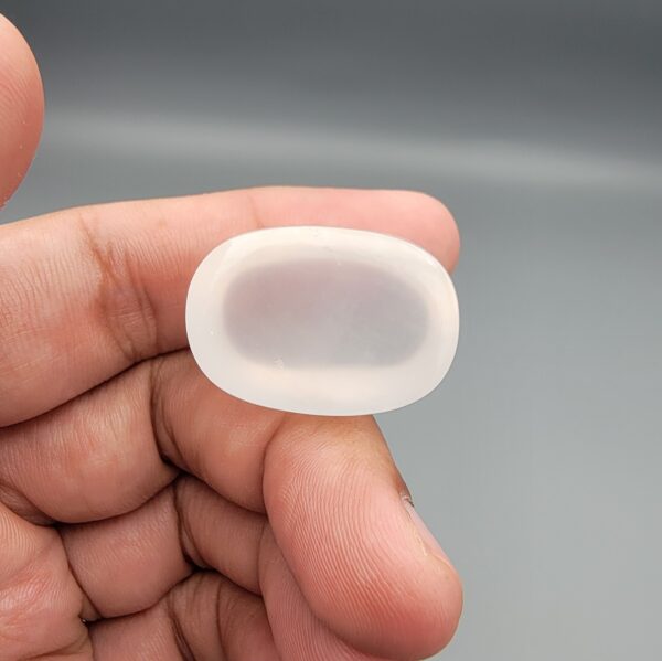 Macro photograph showing a monumental 75.44-carat natural Durr-e-Najaf (Clear Rock Crystal Quartz) cabochon resting on fingertips, featuring a large smooth oval dome, water-clear transparency, and subtle organic internal veil inclusions.
