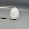 Macro photograph of a 14.40-carat natural Durr-e-Najaf (Clear Rock Crystal Quartz) cabochon held vertically in stainless steel gem tweezers, showcasing a high-domed oval cut, water-clear translucency, and soft internal feather inclusions.