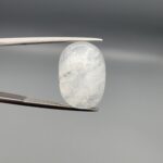 Macro photograph of a 14.40-carat natural Durr-e-Najaf (Clear Rock Crystal Quartz) cabochon held vertically in stainless steel gem tweezers, showcasing a high-domed oval cut, water-clear translucency, and soft internal feather inclusions.