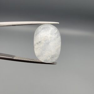 Macro photograph of a 14.40-carat natural Durr-e-Najaf (Clear Rock Crystal Quartz) cabochon held vertically in stainless steel gem tweezers, showcasing a high-domed oval cut, water-clear translucency, and soft internal feather inclusions.