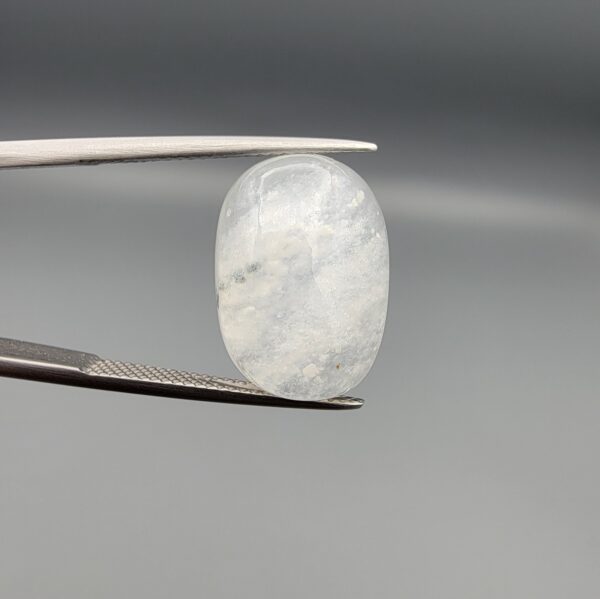 Macro photograph of a 14.40-carat natural Durr-e-Najaf (Clear Rock Crystal Quartz) cabochon held vertically in stainless steel gem tweezers, showcasing a high-domed oval cut, water-clear translucency, and soft internal feather inclusions.