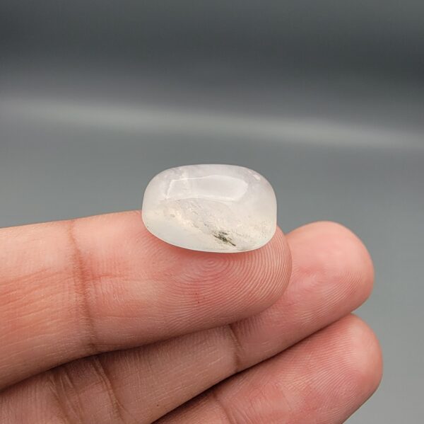 Macro photograph of a 14.40-carat natural Durr-e-Najaf (Clear Rock Crystal Quartz) cabochon held vertically in stainless steel gem tweezers, showcasing a high-domed oval cut, water-clear translucency, and soft internal feather inclusions.