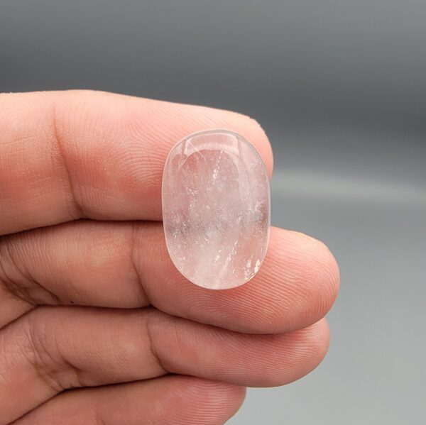 Macro photograph of a 12.29-carat natural Durr-e-Najaf (Clear Rock Crystal Quartz) cabochon resting on fingertips, showcasing an oval polished dome, water-clear translucency, and fine natural internal veil growth patterns.