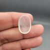 Macro photograph of a 12.29-carat natural Durr-e-Najaf (Clear Rock Crystal Quartz) cabochon resting on fingertips, showcasing an oval polished dome, water-clear translucency, and fine natural internal veil growth patterns.