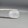 Macro photograph of a 14.05-carat natural Durr-e-Najaf (Clear Rock Crystal Quartz) cabochon held horizontally in stainless steel gem tweezers against a neutral grey background, displaying a smooth oval dome, water-clear transparency, and subtle organic internal veil inclusions.