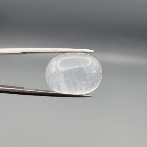 Macro photograph of a 14.05-carat natural Durr-e-Najaf (Clear Rock Crystal Quartz) cabochon held horizontally in stainless steel gem tweezers against a neutral grey background, displaying a smooth oval dome, water-clear transparency, and subtle organic internal veil inclusions.