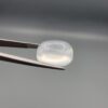 Macro photograph of a 14.05-carat natural Durr-e-Najaf (Clear Rock Crystal Quartz) cabochon held horizontally in stainless steel gem tweezers against a neutral grey background, displaying a smooth oval dome, water-clear transparency, and subtle organic internal veil inclusions.