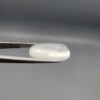 Macro photograph of a 12.48-carat natural Durr-e-Najaf (Clear Rock Crystal Quartz) cabochon held horizontally in stainless steel gem tweezers against a neutral grey background, displaying an elongated smooth oval dome, water-clear transparency, and subtle organic internal veil inclusions.