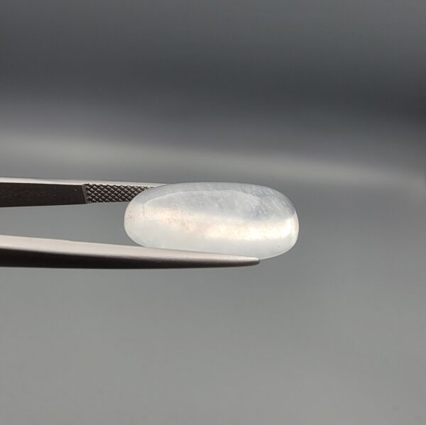 Macro photograph of a 12.48-carat natural Durr-e-Najaf (Clear Rock Crystal Quartz) cabochon held horizontally in stainless steel gem tweezers against a neutral grey background, displaying an elongated smooth oval dome, water-clear transparency, and subtle organic internal veil inclusions.
