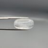 Macro photograph of a 12.48-carat natural Durr-e-Najaf (Clear Rock Crystal Quartz) cabochon held horizontally in stainless steel gem tweezers against a neutral grey background, displaying an elongated smooth oval dome, water-clear transparency, and subtle organic internal veil inclusions.