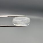 Macro photograph of a 12.48-carat natural Durr-e-Najaf (Clear Rock Crystal Quartz) cabochon held horizontally in stainless steel gem tweezers against a neutral grey background, displaying an elongated smooth oval dome, water-clear transparency, and subtle organic internal veil inclusions.