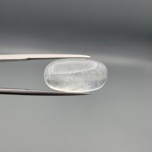 Macro photograph of a 12.48-carat natural Durr-e-Najaf (Clear Rock Crystal Quartz) cabochon held horizontally in stainless steel gem tweezers against a neutral grey background, displaying an elongated smooth oval dome, water-clear transparency, and subtle organic internal veil inclusions.