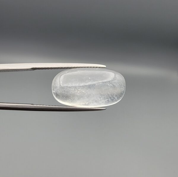 Macro photograph of a 12.48-carat natural Durr-e-Najaf (Clear Rock Crystal Quartz) cabochon held horizontally in stainless steel gem tweezers against a neutral grey background, displaying an elongated smooth oval dome, water-clear transparency, and subtle organic internal veil inclusions.