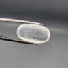 Macro photograph of a 12.48-carat natural Durr-e-Najaf (Clear Rock Crystal Quartz) cabochon held horizontally in stainless steel gem tweezers against a neutral grey background, displaying an elongated smooth oval dome, water-clear transparency, and subtle organic internal veil inclusions.