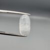 Macro photograph of a 12.09-carat natural Durr-e-Najaf (Clear Rock Crystal Quartz) cabochon held vertically in stainless steel gem tweezers against a neutral grey background, displaying an elongated oval polished dome, water-clear transparency, and subtle organic internal veil inclusions.