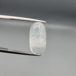 Macro photograph of a 12.09-carat natural Durr-e-Najaf (Clear Rock Crystal Quartz) cabochon held vertically in stainless steel gem tweezers against a neutral grey background, displaying an elongated oval polished dome, water-clear transparency, and subtle organic internal veil inclusions.