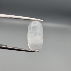 Macro photograph of a 12.09-carat natural Durr-e-Najaf (Clear Rock Crystal Quartz) cabochon held vertically in stainless steel gem tweezers against a neutral grey background, displaying an elongated oval polished dome, water-clear transparency, and subtle organic internal veil inclusions.