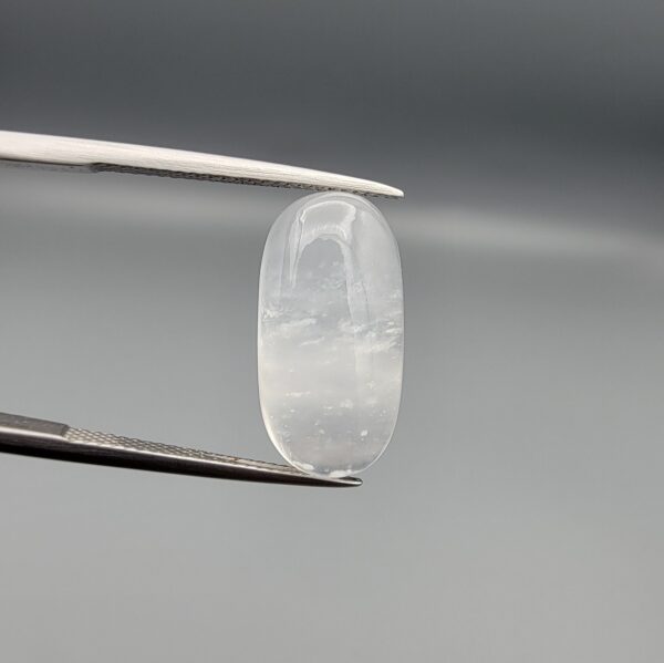 Macro photograph of a 12.09-carat natural Durr-e-Najaf (Clear Rock Crystal Quartz) cabochon held vertically in stainless steel gem tweezers against a neutral grey background, displaying an elongated oval polished dome, water-clear transparency, and subtle organic internal veil inclusions.