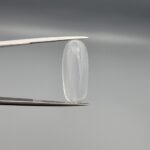 Multi-angle macro photographs of a 9.56-carat natural Durr-e-Najaf (Clear Rock Crystal Quartz) cabochon held vertically in stainless steel gem tweezers and resting on fingers, displaying an elegant slender elongated oval dome, water-clear transparency, and subtle organic internal veil growth.
