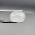 Macro photograph of a 16.91-carat natural Durr-e-Najaf (Clear Rock Crystal Quartz) cabochon held horizontally in stainless steel gem tweezers against a neutral grey background, displaying a smooth oval dome, water-clear transparency, and subtle organic internal veil growth.