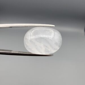 Macro photograph of a 16.91-carat natural Durr-e-Najaf (Clear Rock Crystal Quartz) cabochon held horizontally in stainless steel gem tweezers against a neutral grey background, displaying a smooth oval dome, water-clear transparency, and subtle organic internal veil growth.