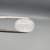 Macro photograph of a 15.34-carat natural Durr-e-Najaf (Clear Rock Crystal Quartz) cabochon held horizontally in stainless steel gem tweezers against a neutral grey background, displaying a smooth oval dome, water-clear transparency, and subtle organic internal veil inclusions.